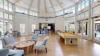 Located at 8800 Pine Forest Rd in Pensacola, Florida, this single-floor community clubhouse offers a generously sized, open-plan living area designed for social gathering and recreation. The centerpiece of the space is a dramatic octagonal vaulted ceiling that rises to impressive heights, adorned with exposed industrial-style ductwork, track lighting, and elegant chandelier fixtures that together create a modern yet welcoming ambiance. Large clerestory windows encircle the upper portion of the room, flooding the interior with natural light and providing views of the surrounding outdoor areas, including what appears to be a pool deck.

The light-toned hardwood flooring runs throughout, complementing the crisp white walls and lending warmth to the expansive layout. A full-size pool table with a bold amber felt surface anchors one side of the room, while a casual lounge area features eclectic patterned accent chairs and a live-edge wood coffee table. Sturdy dark wood structural columns define a secondary gathering zone equipped with a wall-mounted television, adding visual character and architectural interest.

Toward the rear, a stylish blue reception or bar counter serves as a functional focal point, with bar-height seating nearby. Glass doors along the perimeter provide direct access to the outdoor amenity spaces, reinforcing the seamless connection between interior and exterior living.