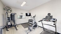 Located at 1102 NE Independence Ave in Lee's Summit, Missouri, this dedicated fitness center offers a well-equipped exercise space designed to support a complete workout routine. The room features vaulted ceilings that lend a sense of openness, while light wood-look flooring and neutral light gray walls create a clean, modern aesthetic throughout.

Natural light flows in generously through multiple windows fitted with blinds and a set of glass-paned French doors, which also provide convenient access to the exterior. A wall-mounted flat-screen television is positioned for easy viewing during workouts, and recessed and overhead lighting ensure the space remains well-lit at all times.

The fitness center is thoughtfully equipped with a variety of cardio machines, including a treadmill, elliptical trainer, and stationary spin bike. A multi-station cable machine and a flat workout bench accommodate strength training needs, while a tiered dumbbell rack stocked with a full set of free weights rounds out the weight training options. Resistance bands are mounted on the wall for additional versatility. Gray exercise mats are placed throughout to protect the flooring and provide cushioning during floor exercises.

A wall mirror enhances the sense of space and allows for form-checking during workouts. Motivational wall lettering adds an encouraging atmosphere. A compact refrigerator and a water dispenser are also on hand for hydration convenience, making this fitness center a fully functional and practical amenity.