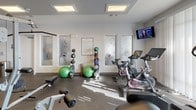 This fitness center offers a comprehensive workout environment designed to support a wide range of exercise routines. The space is laid out across a single floor and is divided into distinct functional zones, allowing users to move seamlessly between cardio, strength, and functional training areas.

The cardio section is equipped with multiple Life Fitness treadmills and stationary cycling bikes, including spin-style cycles, catering to both steady-state and high-intensity workouts. A wall-mounted television is positioned within view of the cardio equipment, enhancing the workout experience. An elliptical trainer and a rowing machine round out the cardio offerings.

The strength training area features a multi-functional cable machine, a weight bench, and an assortment of medicine balls and stability balls neatly organized on racks. This section provides options for both machine-based and functional strength training.

The space is finished with durable rubber flooring throughout, ensuring safety and comfort during exercise. Recessed skylights and flush ceiling fixtures provide bright, even illumination. Frosted glass-paneled doors with decorative etching add a polished, modern aesthetic to the interior. Natural light filters in through large windows, creating an energizing atmosphere.

An accessible restroom is conveniently located within the fitness center, adding to its practicality. Posted fitness center rules ensure a well-maintained and respectful shared environment for all users.