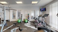 This fitness center offers a comprehensive workout environment designed to support a wide range of exercise routines. The space is laid out across a single floor and is divided into distinct functional zones, allowing users to move seamlessly between cardio, strength, and functional training areas.

The cardio section is equipped with multiple Life Fitness treadmills and stationary cycling bikes, including spin-style cycles, catering to both steady-state and high-intensity workouts. A wall-mounted television is positioned within view of the cardio equipment, enhancing the workout experience. An elliptical trainer and a rowing machine round out the cardio offerings.

The strength training area features a multi-functional cable machine, a weight bench, and an assortment of medicine balls and stability balls neatly organized on racks. This section provides options for both machine-based and functional strength training.

The space is finished with durable rubber flooring throughout, ensuring safety and comfort during exercise. Recessed skylights and flush ceiling fixtures provide bright, even illumination. Frosted glass-paneled doors with decorative etching add a polished, modern aesthetic to the interior. Natural light filters in through large windows, creating an energizing atmosphere.

An accessible restroom is conveniently located within the fitness center, adding to its practicality. Posted fitness center rules ensure a well-maintained and respectful shared environment for all users.