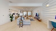 This single-story clubhouse in Jacksonville, Florida, is designed with a cohesive coastal aesthetic, combining functionality with relaxed, resort-inspired style. The interior is unified by light hardwood-style flooring and crisp white shiplap walls accented with thin blue pinstripes, evoking a nautical atmosphere throughout.

The entry foyer welcomes visitors with a herringbone-patterned floor, an elegant chandelier, and a classic pedestal table adorned with fresh florals. From there, the space opens into distinct yet interconnected areas. A spacious lounge features generous sectional seating, a large flat-screen television, and coastal-themed artwork, bathed in natural light from large windows with views of the surrounding outdoor area.

A dedicated seating area with elevated coffered ceilings showcases a woven rattan pendant light, upholstered accent chairs, and a bold mustard-yellow area rug, creating an intimate gathering spot. The business center offers a clean, streamlined workstation with multiple desktop computers, globe pendant lighting, and vibrant aquatic artwork above.

The leasing area includes a white island counter, a beverage refrigerator, coffee station, and wall-mounted displays, providing a polished, welcoming environment. Throughout, the clubhouse balances bright, open spaces with thoughtfully curated furnishings that reflect its Florida coastal setting.