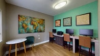 This single-floor business center is thoughtfully designed to serve both productivity and comfort. The room features warm hardwood-style flooring throughout, creating a cohesive and inviting atmosphere. A long granite-topped workstation along one wall accommodates multiple desktop computers and a shared printer, with comfortable tan leather-upholstered high chairs providing ergonomic seating for users. An additional standalone workstation offers a more private computing option.

The space is enlivened by a bold color palette, with a vibrant teal-green accent wall on one side contrasting against warm taupe-gray walls. A striking large-scale world map canvas painting serves as a dramatic focal point, adding an artistic and globally-inspired character to the room. Complementing this, several framed art pieces in earthy and abstract tones are displayed on the teal wall, adding visual depth.

A small marble-topped bistro table paired with a plush emerald-green accent chair provides a casual seating nook, ideal for informal meetings or a quiet break. An oval flush-mount ceiling light ensures even illumination throughout the space. A glass-paneled entry door allows natural light to filter in from the exterior, connecting the interior to the outdoors. A roller shade on the adjacent window offers privacy control as needed.