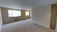 Located at 17 W Chester Pike in Ridley Park, Pennsylvania, this single-floor space offers a clean, functional layout with a welcoming, understated aesthetic. The main living area is generously proportioned, featuring wall-to-wall carpeting in a neutral beige tone that adds warmth and comfort underfoot. Soft, warm-toned walls complement the light palette throughout, creating a bright and airy atmosphere. Multiple windows along the exterior wall flood the room with natural light, enhancing the open feel of the space. A ceiling fan fixture provides both lighting and climate comfort overhead. Practical climate control is well-addressed, with a wall-mounted air conditioning unit and baseboard heating running along the perimeter, ensuring year-round comfort. The clean white trim and baseboards provide a crisp contrast against the warm wall tones, lending the space a tidy, well-maintained appearance. The open floor plan allows for flexible furniture arrangements, making the living area highly adaptable to a variety of uses. Overall, the space presents a straightforward, move-in-ready environment with essential features in place, suited for those seeking a comfortable and functional single-level layout in the Ridley Park area.