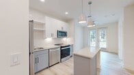 This well-appointed 1-bedroom, 1-bathroom residence offers a thoughtfully designed single-floor layout with a clean, contemporary aesthetic. The open-concept kitchen serves as the heart of the home, showcasing white upper cabinetry, two-tone gray lower cabinets, and a classic white subway tile backsplash. A full suite of stainless steel appliances — including a refrigerator, range, microwave, and dishwasher — pairs with a sleek kitchen island topped with a light quartz surface, illuminated by stylish pendant lighting. Light-toned hardwood flooring flows seamlessly throughout the living areas and bedroom, lending a bright, cohesive feel to the space. A glass-paneled door at the far end of the kitchen area invites natural light deep into the interior while providing access to an outdoor area. The bedroom is a calm, neutral retreat with a ceiling fan, large window with outdoor views, and ample wall space. The bathroom features a white vanity with dark countertops, modern black hardware, and tile flooring, with a dedicated walk-in closet conveniently adjacent. Recessed lighting and a ceiling fan complete the modern, efficient design of this compact yet functional residence.