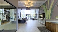 This single-floor space showcases a contemporary, open-plan design that seamlessly blends elegance with functionality. The interior is anchored by a dramatic, sculptural chandelier suspended from a coffered ceiling, serving as an immediate focal point upon entry. Large floor-to-ceiling windows dressed with sheer white drapery flood the space with natural light, creating an airy, inviting atmosphere while offering views of the surrounding outdoor greenery.

The layout is thoughtfully divided into distinct zones. A sleek bar counter runs along one side of the room, lined with modern chrome-based bar stools, making it well-suited for social gatherings or events. On the opposite side, a comfortable lounge area features plush, dark upholstered seating arranged around a central coffee table, ideal for relaxed conversation.

Light-toned hardwood flooring extends throughout, contributing to the bright, cohesive aesthetic. Decorative wall murals add an artistic accent to the space. Glass-paneled doors and walls allow visibility across zones, enhancing the sense of openness. Overall, the space exudes a polished, modern ambiance suited for hosting and community engagement.