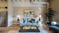 Located on Smallman Street in Pittsburgh's vibrant Strip District, this single-floor unit blends industrial character with contemporary design. The living area immediately draws attention with its soaring ceilings, exposed structural beams, and visible ductwork — hallmarks of the building's converted warehouse heritage. Original brick accent walls add texture and warmth, while large windows invite natural light and offer views of the surrounding urban neighborhood.

The kitchen is well-appointed with white shaker cabinetry, a classic subway tile backsplash, stone-look countertops, and a central island with seating — ideal for casual dining. Pendant lighting and wood-look flooring reinforce the space's modern industrial aesthetic.

The living area provides generous room for comfortable furnishings, with the exposed ceiling elements and brick walls creating a loft-like ambiance. A dedicated sitting nook near the windows offers an intimate space bathed in natural light.

The bathroom features a long vanity with stone-look countertops, white cabinetry with gold hardware, large-format tile flooring, and a full shower-tub combination — all finished in a clean, neutral palette that complements the rest of the unit's design. The overall atmosphere is sophisticated yet approachable, balancing the building's industrial roots with refined, modern touches.