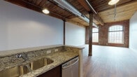 Located at 200 Esten Ave in Pawtucket, Rhode Island, this single-floor mill loft conversion masterfully merges historic industrial character with contemporary upgrades. The open living area is defined by warm hardwood flooring, original heavy-timber ceiling beams, and exposed wood plank ceilings that speak to the building's storied past. Structural columns anchor the generous open floor plan, while exposed ductwork adds an authentic industrial aesthetic. Arched multi-pane windows set within original brick walls flood the space with natural light and lend architectural distinction to the interior.

The kitchen is well-appointed with dark espresso cabinetry, granite countertops, a stainless steel undermount sink, and a full suite of stainless steel appliances including a refrigerator, range, and dishwasher. A peninsula provides additional prep and seating space, seamlessly connecting the kitchen to the living area.

The bathroom features a cohesive, modern design with multi-toned mosaic tile wrapping the walls, large-format porcelain tile flooring, a vessel sink atop a sage green vanity, a wall-mounted mirror, and a full soaking tub with shower combination. Grab bars are thoughtfully integrated throughout, enhancing accessibility and safety.