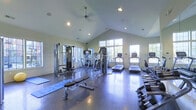 Located at Hayden Lofts in Dublin, Ohio, this well-appointed fitness center offers a comprehensive workout environment designed to accommodate a wide range of exercise routines. The space is defined by its striking vaulted ceilings, which lend an open, airy feel and contribute to a bright and energizing atmosphere. Recessed lighting and a ceiling fan complement the generous natural light that pours in through large, multi-pane windows lining the walls, offering views of the surrounding outdoor greenery.

The flooring throughout is a durable, textured rubber mat surface ideal for both heavy equipment and free-weight use. Strength training is well supported with a cable machine system, a multi-function weight station, an adjustable workout bench, and a dedicated free-weight rack stocked with a full range of dumbbells. A yoga or stretching mat provides space for floor-based exercises and flexibility work.

On the cardio side, the facility includes multiple treadmills, elliptical trainers, and a step machine, several of which are equipped with personal media screens to enhance the workout experience. Wall-mounted mirrors help expand the sense of space while allowing users to monitor their form. The overall layout is thoughtfully organized, providing ample room to move freely between stations, making this a highly functional and inviting fitness facility.