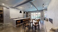 This single-floor communal space showcases a well-considered industrial-modern aesthetic throughout its open layout. Polished concrete floors run continuously across the entire space, complementing exposed silver ductwork overhead that adds an urban, loft-like character. The kitchen area serves as a central hub, featuring a long dark island with a white countertop and bar-height seating for multiple guests, flanked by a striking floor-to-ceiling hexagonal white tile backsplash. Gray cabinetry lines the kitchen wall alongside an undermount sink, refrigerator, and under-counter appliances.

The dining and lounge zones are furnished with black-framed tables and warm wood-toned chairs, creating inviting gathering spots throughout. A recessed linear fireplace is set into a concrete accent wall, accompanied by built-in bench seating nearby. An accent wall clad in reclaimed wood paneling hosts mounted flat-screen televisions, adding a cozy contrast to the industrial surroundings. A counter-height bar along one wall, lined with green metal stools, provides additional casual seating.

Large sliding glass doors open directly to an outdoor courtyard, flooding the interior with natural light and offering views of the surrounding brick architecture. Track lighting and pendant lights provide layered illumination throughout.