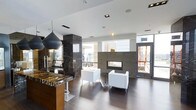 This sophisticated single-floor space showcases a refined contemporary design with an open-concept layout that seamlessly blends functionality and style. The interior is anchored by a striking dark marble column and a richly veined marble-topped bar counter with warm wood cabinetry beneath, creating a bold focal point upon entry. Overhead, a dramatic matte black pendant light adds an artistic contrast to the crisp white ceiling, which features recessed lighting throughout. Dark hardwood floors run across the entire space, grounding the design with warmth and elegance. A sleek, linear gas fireplace set into a smooth grey tile surround serves as the centerpiece of the lounge area, which is furnished with clean-lined white cube chairs and a minimalist side table — ideal for relaxed conversation or reception. Large glass doors along the perimeter allow generous natural light to flood the interior while providing views of and access to the exterior. The combination of polished marble, dark wood tones, white furnishings, and recessed lighting creates a cohesive, upscale ambiance that is both welcoming and architecturally impressive — well suited for a professional, hospitality, or high-end residential common area setting.