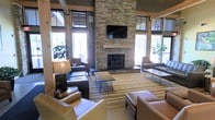 This single-story communal lounge showcases a striking floor-to-ceiling stacked-stone fireplace as its central architectural focal point, complemented by a wall-mounted flat-screen television above a natural wood mantel. The space draws inspiration from rustic lodge design, incorporating warm wood accents, exposed structural beams, and large multi-pane windows that flood the interior with natural light while offering views of surrounding greenery. The generous ceiling height enhances the open, airy feel of the room. Seating is thoughtfully arranged around the fireplace, featuring leather sofas and armchairs in rich brown tones, anchored by a striped, earth-toned area rug with hairpin-leg coffee tables. Additional accent chairs and side tables are positioned near the windows to take advantage of natural light. A decorative table lamp with a woven shade adds a warm, ambient glow. Indoor plants positioned near the windows introduce a natural element, reinforcing the lodge aesthetic. The overall ambiance is inviting and comfortable, balancing rustic charm with modern amenities, making it well-suited as a gathering or relaxation area.
