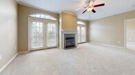 This single-floor residence offers a thoughtfully designed layout with warm neutral tones, crown molding, and plush carpeting throughout the main living areas. The spacious living room is a standout, featuring a tiled fireplace framed by a white mantel, flanked by arched windows and French doors that invite generous natural light and outdoor views. A ceiling fan with a light fixture adds both comfort and character to the room. Adjacent to the living area, an open-concept layout connects seamlessly to a kitchen pass-through, which features rich cherry wood cabinetry, granite countertops, stainless steel appliances including a side-by-side refrigerator, built-in microwave, and electric range, as well as tile flooring. Built-in shelving units near the kitchen entry provide practical display and storage space. The bedrooms are well-proportioned, each with soft carpeting and access to natural light. The primary suite includes a generous walk-in closet with built-in shelving and hanging rods. Two bathrooms serve the residence — one with a tiled tub-shower combination and vanity with granite counters, and another with a glass-enclosed shower and mirrored vanity. Hardwood flooring accents the dining and transitional areas, adding warmth and contrast to the overall design.