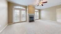 This single-floor residence offers a thoughtfully designed layout with warm neutral tones, crown molding, and plush carpeting throughout the main living areas. The spacious living room is a standout, featuring a tiled fireplace framed by a white mantel, flanked by arched windows and French doors that invite generous natural light and outdoor views. A ceiling fan with a light fixture adds both comfort and character to the room. Adjacent to the living area, an open-concept layout connects seamlessly to a kitchen pass-through, which features rich cherry wood cabinetry, granite countertops, stainless steel appliances including a side-by-side refrigerator, built-in microwave, and electric range, as well as tile flooring. Built-in shelving units near the kitchen entry provide practical display and storage space. The bedrooms are well-proportioned, each with soft carpeting and access to natural light. The primary suite includes a generous walk-in closet with built-in shelving and hanging rods. Two bathrooms serve the residence — one with a tiled tub-shower combination and vanity with granite counters, and another with a glass-enclosed shower and mirrored vanity. Hardwood flooring accents the dining and transitional areas, adding warmth and contrast to the overall design.