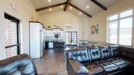 This single-story space is defined by its striking vaulted ceiling adorned with dark wood exposed beams, giving the interior a sense of openness and architectural character. Recessed lighting is integrated throughout the ceiling, providing consistent illumination across the open-plan layout.

The space combines a lounge area, dining zone, and kitchenette within a single flowing floor plan. The lounge area is furnished with large, tufted leather sofas arranged to encourage gatherings and relaxed socializing. Adjacent to the seating area, industrial-style bistro tables with adjustable stools offer casual dining or workstation options.

The kitchenette is equipped with white upper cabinetry, dark lower cabinetry, a stainless steel refrigerator, a dishwasher, and a sink with a window above it. Dark granite-style countertops complement the two-tone cabinetry design.

Rich hardwood-style flooring runs throughout the entire space, tying the zones together cohesively. A set of French doors at the rear allows natural light to filter in and provides access to an exterior area. Neutral warm-toned walls and decorative wall art contribute to an inviting, comfortable ambiance.