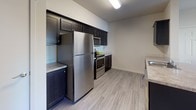 This single-floor unit offers a thoughtfully designed layout that balances comfort and functionality across its key living areas. The kitchen is a standout feature, outfitted with sleek dark cabinetry, stainless steel appliances including a full-size refrigerator, microwave, and range, along with light stone-finish countertops and light wood-look flooring that lends the space a clean, modern aesthetic. A sitting area provides an intimate retreat, furnished with two mid-century-style upholstered armchairs in a warm gray tone, positioned around a minimalist round glass side table with a marble base. A wall-mounted mini-split air conditioning unit ensures year-round climate comfort in this area. The bedroom is a calm, neutral space with soft wall-to-wall carpeting, white blinds filtering natural light through a window, and clean white walls that create a restful atmosphere. The bathroom features tile flooring, a dark wood vanity with a stone-patterned countertop, chrome fixtures, and a shower-tub combination. The overall design of the unit is characterized by neutral tones, practical finishes, and a cohesive layout that maximizes the available space efficiently.