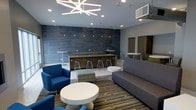 This contemporary single-floor lounge space is thoughtfully designed to encourage both relaxed socializing and casual collaboration. The room is anchored by a sleek built-in bar counter finished in warm wood tones and set against a striking floor-to-ceiling textured slate-blue accent wall, above which a row of pendant lights adds atmospheric illumination. Bar-height stools line the counter, creating an inviting spot for casual conversation or informal meetings.

The seating area is generously appointed with a mix of plush blue armchairs, a large gray sofa, and a striped upholstered ottoman, all arranged around low white round tables atop a bold patterned area rug. This layout encourages flexible use of the space for both intimate conversations and larger group gatherings.

Overhead, a dramatic starburst chandelier serves as a striking focal point, complemented by recessed lighting throughout for a bright, welcoming atmosphere. The neutral gray walls and light flooring provide a clean, modern backdrop that ties the color palette together cohesively. Additional high-top tables along the perimeter offer further options for standing or perching. The overall ambiance is polished yet approachable, reflecting a well-curated modern aesthetic suited to a community or hospitality setting.