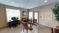 This single-floor space functions as a combined lounge and business center, thoughtfully designed to accommodate both relaxed gathering and productive work. The room is finished with neutral beige walls and wall-to-wall carpet, creating a warm and approachable atmosphere throughout. Recessed lighting is distributed evenly across the ceiling, supplementing the generous natural light that enters through multiple windows dressed with white blinds and sheer curtains. These windows also provide views of the surrounding outdoor greenery, enhancing the room's inviting character.

A central seating area features a round granite-topped table accompanied by upholstered chairs with nailhead trim, positioned atop a patterned area rug. A wooden storage bench with accent pillows is set against one wall, adding both function and charm. A decorative world map and circular framed mirror contribute to the curated aesthetic.

On the opposite side, a dedicated workspace includes a dark wood desk equipped with a computer monitor, task lamp, and potted plant. Nearby, a black chest of drawers supports a printer, offering practical office amenities. A glass-paned interior door leads to an adjoining corridor, and double doors provide additional storage. A classic wall clock and framed artwork complete the well-appointed interior.