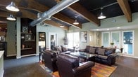 This single-floor space showcases a compelling blend of industrial and rustic design elements that create a distinctive, inviting atmosphere. The ceiling is a standout architectural feature, displaying exposed silver ductwork running alongside dark-painted surfaces and heavy, recessed wood beams that add warmth and texture overhead. Pendant lighting in a classic barn-style finish hangs from the ceiling, casting a focused, ambient glow throughout the space.

The main lounge area is anchored by a generous arrangement of dark brown leather sofas and armchairs, positioned around a glass-topped coffee table. A vibrant, multicolored area rug grounds the seating arrangement and introduces an energetic contrast to the otherwise earthy tones of the room. Wall-mounted artwork and framed pieces add visual interest, while a large mirror near the entry enhances the sense of depth.

Natural light filters in through windows along one wall, brightening the space and providing views of the surrounding exterior. A secondary seating or social area is visible through an open passageway, suggesting a well-connected, open-concept layout. The overall ambiance is bold, comfortable, and well-curated.