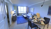 This single-floor space combines a living area and dining space in an open, functional layout. The living area is anchored by a generously sized light-grey sectional sofa, positioned to face a sleek media console along the wall. A bold blue area rug with wave-like patterns adds a striking visual element underfoot, complementing the neutral carpeted flooring throughout. A rectangular coffee table sits at the center, and decorative throw pillows add pops of color to the seating arrangement. Large windows along one wall allow abundant natural light to fill the room, creating a bright and airy atmosphere. Crown molding runs along the ceiling, adding a refined architectural detail to the space. The dining area, situated just adjacent to the living room, features a round dark table paired with modern chairs, set with dishware and glassware. Wall art and a stylish floor lamp contribute to the overall polished aesthetic. The space is designed with a cohesive, contemporary style throughout, making efficient use of the open-plan layout to create a comfortable and visually appealing environment.