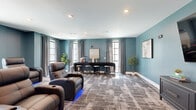 Located at 34 Ramsey Branch Rd in Freeport, FL, this single-floor living space is thoughtfully designed as a premium media and lounge room. The interior is defined by its soothing blue-gray wall color, which lends a calm, contemporary ambiance throughout. Patterned, textured carpeting in neutral earth tones covers the floor, adding warmth and visual interest to the space.

The room is anchored by a large wall-mounted flat-screen television positioned above a sleek, dark gray storage credenza. Arranged in a tiered, theater-style layout, a row of plush leather power recliner chairs — equipped with cup holders and ambient LED accent lighting — provides a true cinematic viewing experience. Framed artwork and sculptural wall pieces add decorative character to the space.

Toward the rear, a dark bar counter with bar-height stools serves as a casual gathering and refreshment area, flanked by tall windows dressed in gray drape curtains that allow natural light to filter in while maintaining privacy. Potted indoor plants and framed art pieces are distributed throughout, contributing to an inviting and polished atmosphere. Recessed ceiling lighting ensures even, functional illumination across the entire room. This well-appointed space is ideal for entertainment and social gatherings.