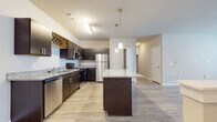 This single-floor residence showcases a thoughtfully designed open-concept layout that seamlessly connects the kitchen, dining, and living areas. The kitchen is a standout feature, equipped with rich dark espresso cabinetry, granite countertops, and a full suite of stainless steel appliances including a refrigerator, range, microwave, and dishwasher. A built-in wine rack adds a distinctive touch above the upper cabinets, while a central granite-topped island with pendant lighting overhead serves as both a functional workspace and a natural gathering point. Light wood-look flooring runs throughout the main living areas, complementing the neutral, light-toned walls and creating a clean, contemporary aesthetic. The living area is bright and airy, benefiting from natural light through windows and a glass door, and features a ceiling fan for added comfort. The bedrooms are finished with plush carpeting and generous windows that invite natural light while maintaining privacy. The bathrooms are well-appointed, with one featuring a full soaking tub and shower combination, and another offering a dark-cabineted vanity with a granite countertop and a large mirror. Track lighting and pendant fixtures throughout the space provide both functional and ambient illumination, contributing to the overall modern and polished feel of this well-designed residence.