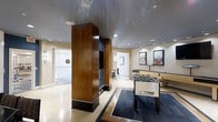 This game room offers a well-appointed recreational space designed for social entertainment. The room features a distinctive design anchored by tall, warm wood-clad columns that add architectural character and visual depth. A dark, reflective ceiling paired with recessed lighting creates a sleek, polished ambiance throughout the space.

At the center of the room, a Tornado foosball table sits atop a geometric area rug with contrasting dark and light tones, serving as a focal point for casual gameplay. Adjacent to it, a shuffleboard table provides an additional gaming option, making the space well-suited for group activities. Framed artwork adorns the walls, contributing to the refined, contemporary aesthetic.

The light-toned flooring contrasts effectively with the darker accents, enhancing the open feel of the room. An adjoining space is visible through a doorway, suggesting additional areas for use. The overall design balances a modern, upscale look with a comfortable, inviting atmosphere ideal for recreation and social gatherings.