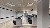 Located at 1001-1003 Easton Rd in Willow Grove, PA, this thoughtfully designed screening room delivers a refined, cinema-quality experience within a compact yet efficient single-floor layout. The space is anchored by multiple rows of plush, high-back leather theater seats arranged on a tiered platform, ensuring clear sightlines to a large wall-mounted projection screen at the front of the room. A ceiling-mounted projector provides a professional-grade viewing setup, while vertical slatted acoustic wall panels enhance sound quality and contribute to the space's polished aesthetic. The flooring transitions between distressed wood-look vinyl planks and a vibrant teal area rug, adding warmth and visual contrast to the otherwise neutral gray palette. A decorative coffered ceiling detail with metallic inlays draws the eye near the entry area, complementing the standard drop tile ceiling throughout the rest of the space. Along one side, a built-in wet bar with dark countertops, open shelving, and a sink provides convenient refreshment service. Adjacent to the bar, a glass partition offers views into a neighboring area, while clearly marked exit doors ensure straightforward egress. Recessed and surface lighting throughout maintains a well-lit, welcoming ambiance suited for presentations, screenings, or private events.