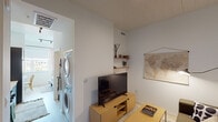 This single-floor unit is thoughtfully designed with a modern, minimalist aesthetic that emphasizes clean lines, bright white walls, and light wood flooring throughout. The living area serves as the heart of the space, featuring built-in floating shelves, a wall-mounted map as décor, and a media console with a flat-screen TV. A decorative coffee table adds a functional touch to the seating area, while a patterned area rug grounds the space. The kitchen is a standout feature, outfitted with sleek dark cabinetry, white countertops, a white tile backsplash, and stainless steel appliances including a gas range with oven, over-the-range microwave, and dishwasher. The bedroom is bright and inviting, benefiting from generous natural light through wide windows that offer open outdoor views. Light wood floors, a full-size bed, a wooden nightstand, and a curated gallery wall lend the room a warm, personal character. A large mirror with a dark frame enhances the sense of space. A dedicated laundry area with a stacked washer and dryer unit adds practical convenience. The unit is connected by a central hallway punctuated by a bold dark-painted door, tying together the modern design palette.