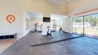 This single-story fitness center is designed to accommodate a comprehensive workout experience within an open, well-organized layout. The space is divided into distinct functional zones, seamlessly connected through wide architectural openings framed by clean white columns and crown molding details.

The cardio zone is anchored by floor-to-ceiling windows that flood the area with natural light while offering views of the surrounding landscape and pool area. Equipment includes treadmills, an elliptical, a stationary bike, and a Concept2 rowing machine. A wall-mounted flat-screen TV and a ceiling fan add comfort and entertainment for cardio sessions. Motivational phrases such as "Harder. Better. Faster. Stronger." and "Hustle for Muscle" are prominently displayed on the walls.

The strength and functional training area features a power rack with barbells, medicine ball storage, battle ropes, and additional free weights. A large mirror maximizes the sense of space and supports form monitoring during workouts. Wall-mounted mini-split HVAC units ensure climate control throughout.

Durable dark rubber tile flooring covers the entire workout area, transitioning to hardwood near the entrance. Recessed lighting combined with abundant natural light creates a bright, energizing atmosphere throughout the facility.
