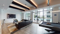Located on NW 11th Ave in Miami, FL, this single-floor amenity lounge showcases a sleek, contemporary design tailored for social gatherings and leisure. The open-plan layout is anchored by warm wood-look flooring and a striking ceiling treatment featuring bold dark exposed beams with integrated ambient uplighting, adding depth and architectural character to the space.

Expansive floor-to-ceiling windows and glass walls span the length of the room, flooding the interior with abundant natural light and offering unobstructed views of the resort-style outdoor pool and lounge deck beyond. This seamless connection between indoor and outdoor spaces creates an airy, resort-like atmosphere.

The lounge area is furnished with generous tufted leather sofas arranged over layered area rugs, providing a comfortable and sophisticated setting. A full-size billiards table serves as a central entertainment focal point, complemented by a wall-mounted TV and a dark, built-in shelving unit displaying curated décor and books. A compact wet bar with stainless steel appliances and white cabinetry offers convenient refreshment access. An adjacent mirrored room provides additional private space. Together, these elements create a well-appointed, multifunctional amenity space that reflects Miami's vibrant, upscale lifestyle.