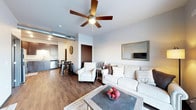 This thoughtfully designed single-floor space offers an open-concept layout that seamlessly connects the living area, dining space, and kitchen. The living room is anchored by a comfortable neutral-toned sofa and armchair arrangement centered around a rustic coffee table, creating an inviting atmosphere for relaxation and entertaining. A ceiling fan with integrated lighting overhead adds both function and a casual modern touch to the space. Exposed ductwork along the ceiling lends an industrial character that complements the warm, hardwood-style flooring running throughout. The kitchen, visible from the living area, features dark cabinetry contrasted against light countertops and is equipped with stainless steel appliances, including a dishwasher. A compact dining area with a table and chairs sits adjacent to the kitchen, making everyday meals convenient and accessible. A framed mirror on the living room wall enhances the sense of openness, while neutral wall tones throughout contribute to a clean, cohesive aesthetic. The overall ambiance is warm and contemporary, blending modern finishes with comfortable, livable design elements that make the space both functional and welcoming.