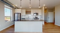 This contemporary single-floor unit is thoughtfully designed with a clean, modern aesthetic throughout. The open-concept kitchen serves as the centerpiece, showcasing crisp white shaker cabinetry, a classic white subway tile backsplash, and dark quartz countertops that extend across a generously sized island. Three globe pendant lights hang above the island, adding a stylish focal point, while recessed lighting and stainless steel appliances — including a refrigerator, electric range, and microwave — complete the well-appointed space. Light hardwood flooring flows seamlessly from the kitchen into the living area, which is bathed in natural light from large windows and a sliding glass door that opens to a private balcony offering sweeping views of the surrounding cityscape and tree canopy. The bedrooms are carpeted for comfort, each featuring windows with exterior views and exposed ductwork that lends a subtle industrial character. The unit includes two bathrooms: one with a tub-shower combination and single vanity, and a second featuring a dual-sink vanity with dark hardware and quartz countertops, offering ample storage. Throughout the unit, consistent use of black hardware accents, neutral tones, and quality finishes create a cohesive and refined living environment.