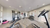 This fitness center is a thoughtfully designed exercise space that offers a comprehensive range of workout equipment within a single, efficiently utilized room. The space features a herringbone-patterned carpet in neutral blue and tan tones, lending a modern aesthetic that sets it apart from a typical gym environment.

The cardio zone is well-appointed with a treadmill, elliptical trainer, rowing machine, and a recumbent stepper, providing diverse options for cardiovascular workouts. Strength training is equally addressed, with a multi-function cable machine, a weight bench, a punching bag on a freestanding stand, and a rack of dumbbells visible in the mirrored wall reflections.

A full wall of white-framed mirrors runs along one side of the room, creating a sense of expanded space while allowing users to monitor their form during exercise. A wall-mounted flat-screen television is positioned prominently above the mirrors, enhancing the workout experience. Motivational text art on the wall adds an energizing atmosphere to the space.

Natural light fills the room through multiple white-framed windows that offer views of lush outdoor greenery, contributing to a bright and welcoming environment. Recessed lighting supplements the natural light, ensuring the space remains well-lit at all times. A glass-panel door provides direct outdoor access. A water fountain is also available for convenience.