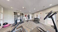 This fitness center is a thoughtfully designed exercise space that offers a comprehensive range of workout equipment within a single, efficiently utilized room. The space features a herringbone-patterned carpet in neutral blue and tan tones, lending a modern aesthetic that sets it apart from a typical gym environment.

The cardio zone is well-appointed with a treadmill, elliptical trainer, rowing machine, and a recumbent stepper, providing diverse options for cardiovascular workouts. Strength training is equally addressed, with a multi-function cable machine, a weight bench, a punching bag on a freestanding stand, and a rack of dumbbells visible in the mirrored wall reflections.

A full wall of white-framed mirrors runs along one side of the room, creating a sense of expanded space while allowing users to monitor their form during exercise. A wall-mounted flat-screen television is positioned prominently above the mirrors, enhancing the workout experience. Motivational text art on the wall adds an energizing atmosphere to the space.

Natural light fills the room through multiple white-framed windows that offer views of lush outdoor greenery, contributing to a bright and welcoming environment. Recessed lighting supplements the natural light, ensuring the space remains well-lit at all times. A glass-panel door provides direct outdoor access. A water fountain is also available for convenience.