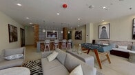 This single-floor amenity space combines stylish interior gathering areas with a well-appointed outdoor courtyard. The interior lounge and game room is a generously sized, open area anchored by a foosball table at its center, surrounded by comfortable seating arrangements including a large sectional sofa, built-in bench seating with decorative pillows, and accent side tables. Bold black-and-white artwork adorns the walls, and a wall-mounted television adds to the entertainment options. Recessed lighting and pendant fixtures illuminate the space throughout. Adjacent to the lounge is a catering-style kitchen featuring warm wood cabinetry, light countertops, and a striking accent wall clad in rustic brick tile alongside a decorative patterned tile backsplash. A bar counter with barstool seating provides an additional social hub. The exterior of the building showcases a limestone facade with arched entryways and stainless steel railings leading into the amenity level. The courtyard is a standout feature, offering a spacious open-air setting with manicured greenery, gravel landscaping, and a wood pergola fitted with ceiling fans. Multiple outdoor dining and lounge seating arrangements are spread across the tiled and grassy courtyard areas, creating a relaxed and social outdoor atmosphere.