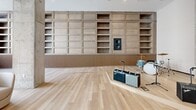 Located at 915 Division Street in Nashville's vibrant Gulch district, this single-floor space is thoughtfully designed as a music-inspired lounge and gathering area. The open layout offers a generous, flowing footprint that supports both intimate conversation and live performance. Light hardwood flooring runs throughout, lending warmth and continuity across the entire space.

The main living area is anchored by floor-to-ceiling windows along multiple walls, framing sweeping views of the surrounding Nashville skyline and flooding the interior with abundant natural light. Exposed concrete columns add an industrial character, contrasting elegantly with the refined finishes throughout.

A dedicated performance zone features a raised stage area equipped for live music, complete with a grand piano and guitar amplifiers, complemented by an expansive wall of built-in wood shelving with an integrated speaker system. A series of illuminated wall niches display a curated collection of guitars, celebrating Nashville's musical heritage.

The lounge seating area is furnished with modern, low-profile sofas and rounded upholstered chairs arranged around circular coffee tables, creating a relaxed yet sophisticated atmosphere. Acoustic ceiling panels are visible overhead, ensuring the space is optimized for sound quality. Overall, the design balances functionality with a distinctly music-centric aesthetic.