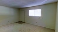 This single-floor space presents a straightforward and functional layout. The main room is characterized by neutral, light-colored walls that create a clean and understated atmosphere. The flooring throughout is covered in wall-to-wall carpet in a muted, beige tone, providing a soft underfoot feel and contributing to the room's quiet, subdued aesthetic. A standard interior door is positioned along one wall, offering access to an adjoining area. A window fitted with horizontal blinds allows natural light to enter the space, brightening the interior while also offering privacy and light control. The ceiling is smooth and painted white, maintaining the room's simple, no-frills design. Electrical outlets and a light switch are visibly positioned along the walls, reflecting a practical and utilitarian approach to the space's design. Overall, the space is modest and unadorned, presenting a blank canvas that is well-suited for a variety of uses. The open, uncluttered nature of the room allows for flexible furniture arrangements and personalization to meet the needs of its occupants.