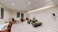 Located at 7971 S Sixth St in Oak Creek, Wisconsin, this thoughtfully designed single-floor space seamlessly blends a comfortable lounge area with a productive workspace. The room is finished with light wood-look flooring and warm beige walls, creating a clean and welcoming atmosphere throughout.

The lounge area features green leather seating, including a sofa and accent chairs, arranged atop a circular patterned area rug. A tan leather mid-century chair adds a complementary design touch, while open white shelving displays decorative accents and greenery. A distinctive tray ceiling with integrated ambient lighting runs along one wall, adding architectural depth and a soft, layered glow to the space.

The workspace area accommodates two white adjustable-height desks, each paired with cognac leather rolling chairs, offering a functional and stylish environment for productivity. A series of bold black wall art panels with abstract designs are displayed prominently across the walls, tying together the room's modern aesthetic.

Recessed ceiling lighting throughout ensures the space is well-lit, while HVAC vents are neatly integrated into the ceiling. A white interior door and a contrasting dark-framed glass entry provide clear transitions within the space. Overall, the room strikes a refined balance between comfort and function, making it well-suited for both collaborative and focused use.