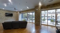 This single-floor cabana offers a well-appointed gathering space that seamlessly blends indoor comfort with outdoor accessibility. The interior is defined by striking natural stone masonry columns and accent walls that lend a warm, resort-like character to the space. Polished concrete flooring in a rich, warm tone runs throughout, providing a durable yet visually appealing surface that anchors the room's aesthetic.

Expansive floor-to-ceiling sliding glass doors span much of the exterior-facing walls, flooding the interior with abundant natural light and providing wide, unobstructed views of the outdoor patio area. The patio beyond features shaded seating arrangements and mature palm trees, reinforcing the relaxed, cabana atmosphere. The seamless transition between the interior lounge area and the outdoor space makes this cabana ideal for social gatherings and leisure.

Ceiling fans are mounted overhead, ensuring comfortable airflow throughout the space. Recessed lighting supplements the natural light for evening use. A comfortable sectional sofa anchors the lounge area, offering ample seating for guests. The overall ambiance is casual yet refined, combining natural materials with modern finishes to create an inviting, functional retreat suited for both relaxation and entertainment.