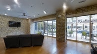 This single-floor cabana offers a well-appointed gathering space that seamlessly blends indoor comfort with outdoor accessibility. The interior is defined by striking natural stone masonry columns and accent walls that lend a warm, resort-like character to the space. Polished concrete flooring in a rich, warm tone runs throughout, providing a durable yet visually appealing surface that anchors the room's aesthetic.

Expansive floor-to-ceiling sliding glass doors span much of the exterior-facing walls, flooding the interior with abundant natural light and providing wide, unobstructed views of the outdoor patio area. The patio beyond features shaded seating arrangements and mature palm trees, reinforcing the relaxed, cabana atmosphere. The seamless transition between the interior lounge area and the outdoor space makes this cabana ideal for social gatherings and leisure.

Ceiling fans are mounted overhead, ensuring comfortable airflow throughout the space. Recessed lighting supplements the natural light for evening use. A comfortable sectional sofa anchors the lounge area, offering ample seating for guests. The overall ambiance is casual yet refined, combining natural materials with modern finishes to create an inviting, functional retreat suited for both relaxation and entertainment.