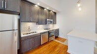 Located in the heart of Richmond, VA on North Ninth Street, this single-floor urban residence blends historic character with modern updates. Rich, original hardwood floors flow throughout the open living areas, adding warmth and a sense of authentic craftsmanship to the space.

The living area is defined by oversized, black-framed windows that frame sweeping views of downtown Richmond's historic skyline. Natural light pours in generously, highlighting the bright white walls and soaring ceilings equipped with a ceiling fan and recessed lighting. Pendant lights accent the transition between the living area and kitchen.

The kitchen is thoughtfully designed with dark espresso cabinetry, light quartz countertops, a textured subway tile backsplash, and a full suite of stainless steel appliances including an electric range, built-in microwave, and dishwasher. Additional counter and cabinet space lines both sides of the kitchen, maximizing storage and functionality.

The bathroom features a clean, contemporary aesthetic with light wood-look tile flooring, white shaker cabinetry, granite countertops, a large vanity mirror, and a tiled walk-in shower with a classic white subway tile surround. A sizable window in the bathroom invites natural light while maintaining privacy. The overall ambiance is a refined blend of urban history and modern comfort.