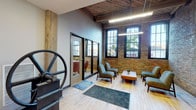 This single-floor industrial loft conversion masterfully blends historic character with modern living. The space retains its original exposed brick walls, rich wood-beamed ceilings, and large multi-pane arched windows that bathe the interior in abundant natural light and frame views of surrounding greenery. Polished light wood flooring runs throughout, complementing the warm tones of the brick and timber.

The entry area makes an immediate impression, featuring a preserved industrial machine — a large cast-iron wheel mechanism — that serves as a striking architectural artifact and nod to the building's heritage. Glass-panel interior doors and sleek pendant lighting enhance the modern sensibility layered atop the industrial bones.

The open-plan living and dining area is generously proportioned, with the kitchen positioned along one wall. Warm wood cabinetry pairs with stainless steel appliances and a clean white countertop, offering a fully functional and stylish cooking space. Exposed ductwork runs along the ceiling, reinforcing the loft's industrial aesthetic.

The living area is anchored by the exposed brick and tall windows, creating an intimate yet airy atmosphere. A bedroom area features clean, neutral walls that provide a calm contrast to the textured brick found elsewhere. Throughout, the design balances raw industrial elements with contemporary finishes, resulting in a cohesive and inviting space full of character.