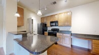 This single-floor residence offers a thoughtfully designed layout that balances functionality and comfort. The kitchen serves as the heart of the home, showcasing rich dark granite countertops, warm honey-toned wood cabinetry, and a full suite of black appliances including a refrigerator, range, and built-in microwave. A curved kitchen island with an integrated sink provides generous prep space and doubles as a casual dining area, while track and pendant lighting illuminate the space effectively. Hardwood-style flooring flows throughout the kitchen and main living areas, lending a cohesive, polished look. A dedicated pantry with a glass-panel door adds convenient storage just steps from the cooking area. The bedroom offers a calm retreat, featuring plush neutral carpet, a ceiling fan with light fixture, and a window with blinds that invites natural light. The bathroom is clean and classic, with tiled walls surrounding a full soaking tub and shower combination, complemented by a granite-topped vanity. A dedicated laundry area accommodates a full-sized washer and dryer side by side, adding everyday convenience. The overall layout, visible through the dollhouse view, presents an efficient and well-connected floor plan suited for comfortable single-floor living.