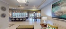 This single-floor amenity space is designed to serve as a well-rounded communal hub, offering a variety of functional zones within an open, cohesive layout. The interior design blends contemporary and transitional styles, featuring a neutral palette of warm grays and beige accented by bold olive-green ceiling details and teal decorative touches throughout.

The main gathering area includes a curved, dark wood bar counter with a white quartz surface, illuminated by a row of crystal pendant lights overhead. Adjacent to the bar, a dedicated business center provides computer workstations and a mounted flat-screen television. A separate lounge zone offers comfortable seating arrangements, including a neutral upholstered sofa, accent chairs in yellow and teal, layered area rugs, and curated wall art, creating an inviting atmosphere for relaxation.

The game room features a full-sized pool table positioned near large windows that line the exterior wall, flooding the space with natural light filtered through white horizontal blinds and sheer teal-accented drapes. A dining area with a glass-top table and acrylic ghost chairs adds a modern, airy touch, while a kitchenette with stainless steel appliances provides convenient access to refreshments. Light tile flooring runs throughout, contributing to the clean, bright overall ambiance.
