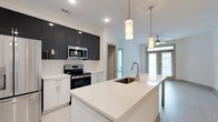 This single-floor space showcases a thoughtfully designed open-concept layout that connects the kitchen and living areas in a clean, contemporary style. The kitchen serves as a focal point, featuring a striking two-tone cabinetry scheme with dark upper cabinets contrasting against crisp white lower cabinets. A generously sized island with a white quartz countertop anchors the space, equipped with an undermount stainless steel sink and pendant lighting suspended above, adding both functionality and visual appeal. Stainless steel appliances, including a range and built-in microwave, complement the modern aesthetic. Recessed lighting and light-toned flooring contribute to a bright, airy atmosphere throughout. The kitchen transitions naturally into the living area, where a ceiling fan and large windows allow abundant natural light to fill the space, creating a welcoming and open ambiance. A glimpse of a walk-in storage or closet space is visible in the background, suggesting practical storage solutions within the unit. The overall design balances modern aesthetics with everyday functionality, making it an inviting and well-appointed living space.