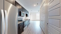 Located at 6001 S Congress Ave in Austin, Texas, this single-story unit offers a clean, contemporary design with consistent finishes throughout. The kitchen is a standout feature, showcasing rich dark cabinetry, granite countertops, a geometric tile backsplash, and a full suite of stainless steel appliances including a refrigerator, electric range, microwave, and dishwasher. Light wood-look flooring runs throughout the main living areas, lending a bright and cohesive feel to the space. The open living area is generously sized, bathed in natural light from large windows fitted with blinds, and equipped with a ceiling fan for added comfort. A door from the living area provides additional access and light. The bedroom continues the light flooring aesthetic, with large windows that fill the room with natural light, and features plush carpeting along with access to a dedicated walk-in closet outfitted with shelving and hanging rods. The hallway offers additional storage with multiple closet doors. The bathroom is thoughtfully finished with tile flooring, a dark vanity with granite countertop, a tiled tub-shower combination, and clean white walls, creating a functional and polished space. Overall, the unit presents a modern, move-in-ready environment with quality finishes and practical storage solutions throughout.
