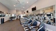 Located at 1564 McNaughten Rd in Columbus, Ohio, this single-floor fitness center is thoughtfully designed to support a comprehensive workout experience. The main exercise area features vaulted ceilings with ceiling fans and recessed lighting, creating a bright and airy atmosphere. Durable rubber flooring spans the space, providing both comfort and functionality underfoot.

The cardio zone is generously outfitted with a row of treadmills, elliptical machines, and upright cycling bikes, all positioned along mirrored walls that enhance the sense of openness. Multiple wall-mounted flat-screen televisions are positioned throughout for entertainment during workouts. A free weights section includes a full dumbbell rack with a range of weights alongside a flat workout bench, catering to strength training needs.

A distinct glass-enclosed studio area, separated by large black-framed glass panels and doors, offers a dedicated space for group fitness or personal training sessions. This private studio is furnished with stability balls, resistance equipment, and a cable machine, making it ideal for functional and flexibility training. Natural light filters in through windows, adding to the welcoming environment. Motivational fitness photography adorns the walls throughout, reinforcing the space's active and focused ambiance.