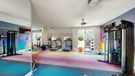 This fitness center is a thoughtfully designed workout facility that combines energetic aesthetics with a comprehensive range of exercise equipment. The space is defined by its striking geometric accent wall, which features a bold mosaic of purple, blue, yellow, and pink polygons, creating a dynamic and motivating atmosphere. Complementing this visual centerpiece, the floor is covered in a vibrant multi-toned carpet that transitions through shades of blue, pink, orange, and yellow, adding to the space's lively character.

The cardio zone is well-appointed with multiple treadmills and elliptical machines, positioned near large windows that invite natural light and provide views of the surrounding greenery. The strength training area houses an Inspire multi-functional cable machine, a dedicated dumbbell rack with a full range of free weights, an adjustable workout bench, and additional resistance machines for a variety of targeted exercises. A dedicated stretching and flexibility area features yoga mats, stability balls, and medicine balls for functional training.

A full-length mirror along one wall enhances the sense of space, while a ceiling fan ensures comfortable air circulation throughout workouts. Recessed lighting provides even, consistent illumination. A wall-mounted television adds entertainment options, and the direct exterior door offers convenient access in and out of the facility.
