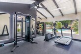 Located in Pleasanton, CA, this well-equipped fitness center is housed in a charming single-room structure defined by its striking vaulted ceiling with bold, dark exposed beams set against white shiplap planking. The soaring ceiling height lends an open, airy feel that belies the room's compact footprint. Large windows and a set of French doors flood the space with natural light while offering pleasant views of an adjacent outdoor pool and surrounding greenery, creating an inviting workout environment.

The fitness center is thoughtfully organized to maximize functionality. Cardio equipment, including a treadmill, elliptical trainer, and rowing-style machine, lines one side of the room near the expansive windows overlooking the pool. On the opposite end, a multi-functional cable machine and a full-length LED-backlit mirror provide a dedicated strength training zone. A dumbbell rack stocked with a complete range of free weights, an adjustable workout bench, and a medicine ball storage rack round out the strength training offerings.

Underfoot, durable gray carpet provides comfortable, low-impact flooring throughout. A ceiling fan ensures air circulation, while a wall-mounted HVAC unit maintains year-round climate control. The clean, neutral color palette of white walls and dark accents gives the space a modern, professional aesthetic suited for a focused and effective workout experience.