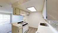 This single-floor space presents a thoughtfully arranged kitchen that emphasizes cleanliness and functionality. The kitchen is outfitted with white upper and lower cabinetry featuring black hardware, offering ample storage for everyday essentials. A white gas range with multiple burners serves as the central cooking appliance, complemented by an overhead range hood for ventilation. White laminate countertops run along both sides of the kitchen, maximizing prep space, while a stainless steel double-basin sink is conveniently positioned along one wall. A flush skylight panel in the ceiling provides bright, diffused overhead lighting, creating a well-lit and airy atmosphere throughout the kitchen. The flooring consists of light wood-tone vinyl planks that lend warmth and are easy to maintain. The walls are painted a neutral off-white, contributing to the space's clean and open feel. The overall layout is efficient and galley-inspired, making practical use of the available space while maintaining a tidy, welcoming environment suitable for everyday cooking and meal preparation.